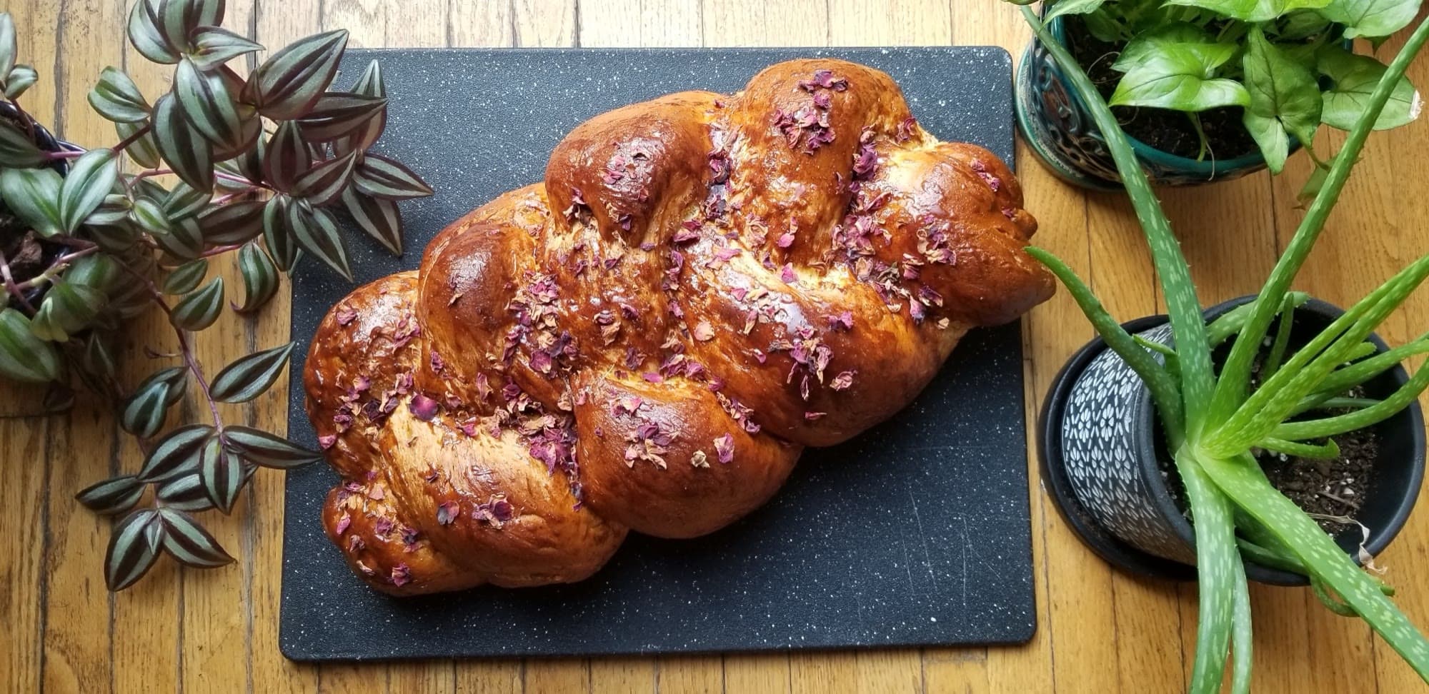 A braided challah topped with rose petals on a dark serving board.