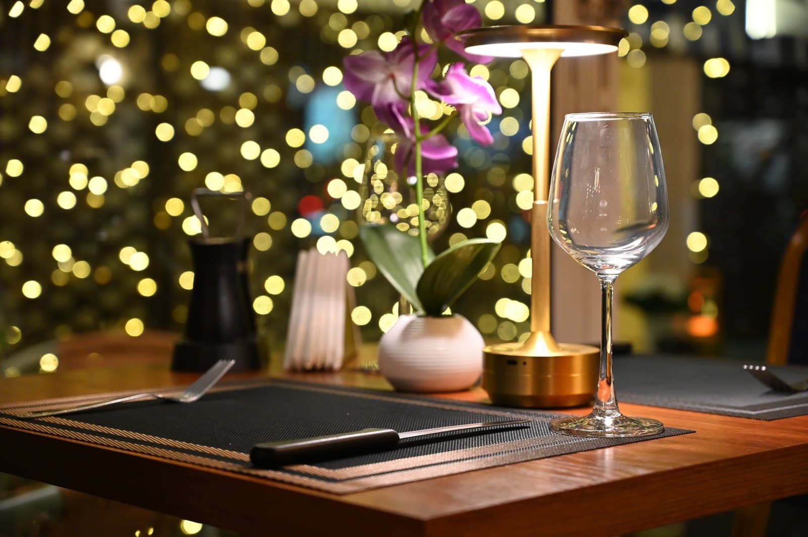 A candlelit restaurant table for two with an orchid, a wine glass, and warm bokeh lights behind.